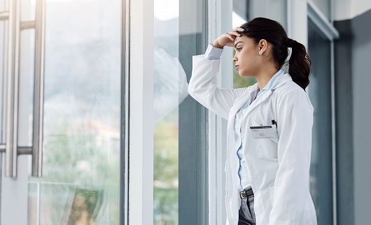 Shot of a young female doctor looking stressed out while standing at a window in a hospital